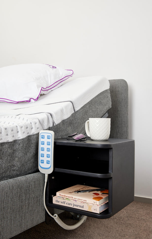 The bedside shelf unit is shown attached to a grey upholstered Icare bed. It holds a white coffee mug and eyeglasses on top, while the lower shelves store a pen and books (including "The Self-Care Journal").