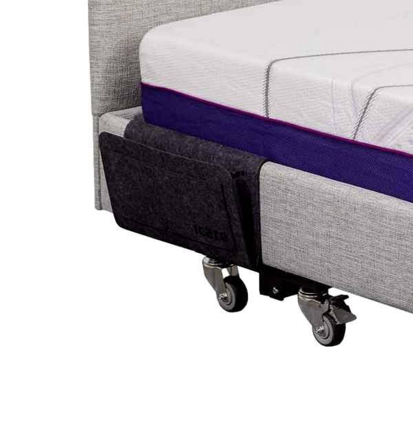 An angled view view of an Icare adjustable bed with a charcoal grey felt bedside pouch attached to the upholstered side surround. The pouch is slim-line and hangs flat against the side of the bed, positioned near the head for easy access to personal items.