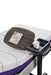 An Icare over bed table positioned over a bed. The table has a wood-grain top with a self-care journal resting on it, and a fixed gray side tray holding a white mug.
