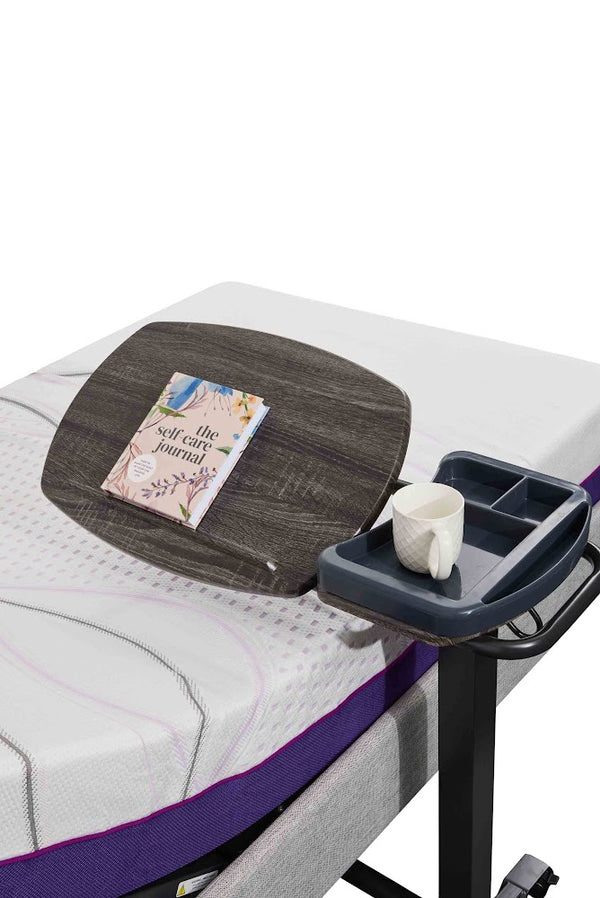 An Icare over bed table positioned over a bed. The table has a wood-grain top with a self-care journal resting on it, and a fixed gray side tray holding a white mug.