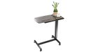 A studio shot of the Icare over bed table isolated on a white background, featuring a wood-grain top, a side tray with a plant and a mug, and a black metal frame with wheels.