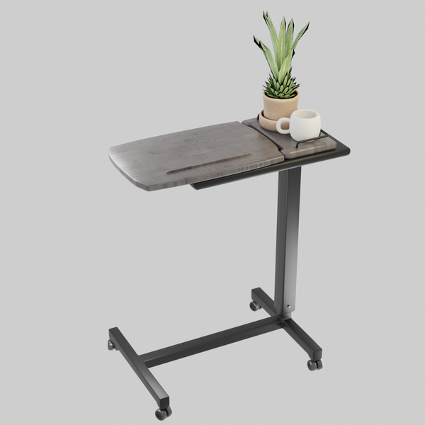 A studio shot of an Icare over bed table on a light gray background. The table features a wood-grain tilting top and a small side tray holding a small potted plant and a white mug. It has a black metal frame with caster wheels.