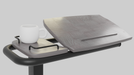 A close-up view of the Icare over bed table top, showing the wood-grain tilting surface slightly elevated and a gray side tray holding a white mug. The view is from the side, emphasizing the tilting mechanism.