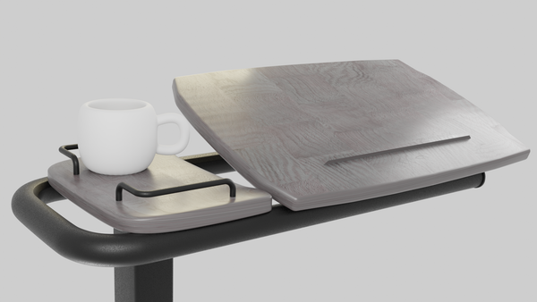 A close-up view of the Icare over bed table top, showing the wood-grain tilting surface slightly elevated and a gray side tray holding a white mug. The view is from the side, emphasizing the tilting mechanism.