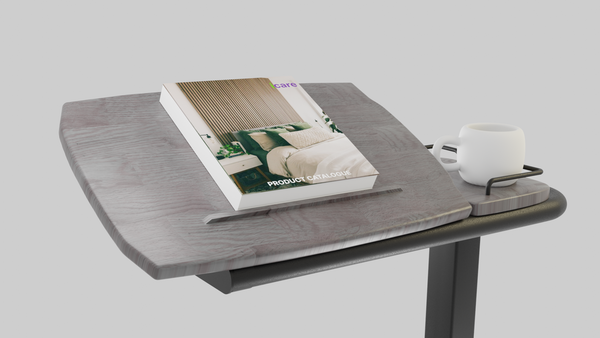 A view of the Icare over-bed table top with a product catalogue resting on the wood-grain surface, which is tilted for reading. A white mug sits on the adjacent fixed side tray.