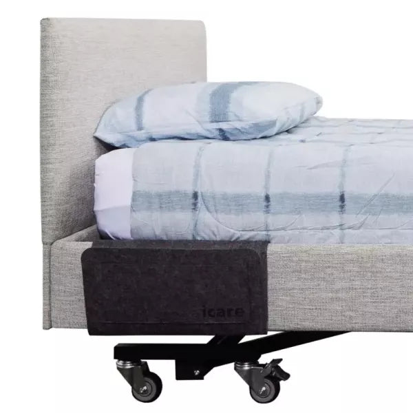 A side profile view of an Icare adjustable bed with a charcoal grey felt bedside pouch attached to the upholstered side surround. The bed is neatly made with a grey textured headboard and blue-and-white checkered bedding. The pouch is slim-line and hangs flat against the side of the bed, positioned near the head for easy access to personal items.