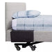 A side profile view of an Icare adjustable bed with a charcoal grey felt bedside pouch attached to the upholstered side surround. The bed is neatly made with a grey textured headboard and blue-and-white checkered bedding. The pouch is slim-line and hangs flat against the side of the bed, positioned near the head for easy access to personal items.