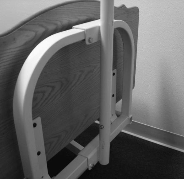 Close-up, black and white image of the Joerns metal trapeze mounting adapter bracket bolted onto the headboard or frame of a hospital bed. The wood-grain headboard is visible behind the adapter.