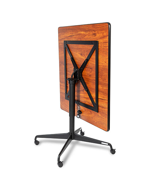 Square tilt top dining table tilted view displaying Biltmore Cherry wood grain surface with black folding base and wheel casters