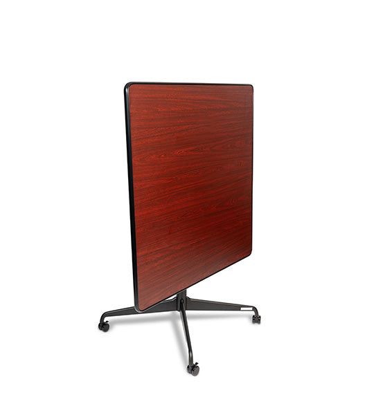 Square tilt top dining table in vertical storage position with Mahogany wood grain top and black metal base with wheel casters