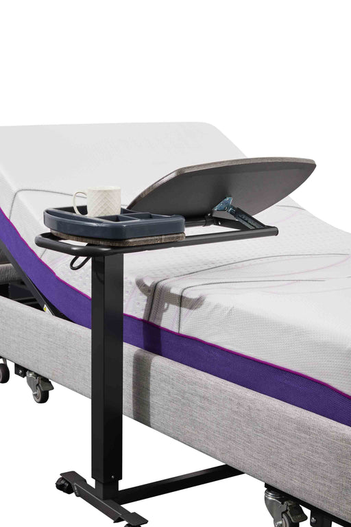 An Icare over bed table positioned over a bed. The table has a dark wood-grain top tilted upwards, with a dark gray organizer tray attached to the side holding a white mug.