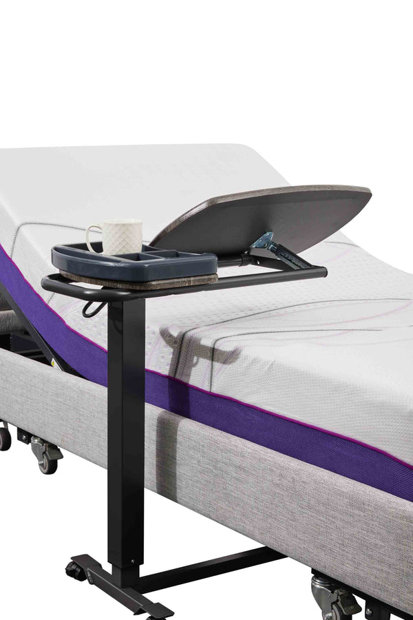 An Icare over bed table positioned over a bed. The table has a dark wood-grain top tilted upwards, with a dark gray organizer tray attached to the side holding a white mug.