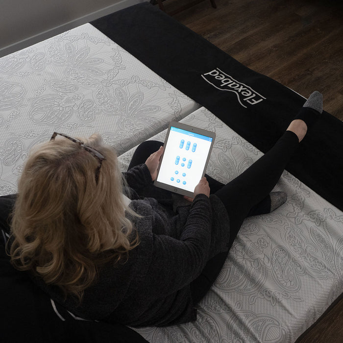 A top-down view of a woman sitting on one side of a Dual Flexabed Premier adjustable bed. She is using a tablet to adjust the bed, with the screen showing multiple customizable settings.