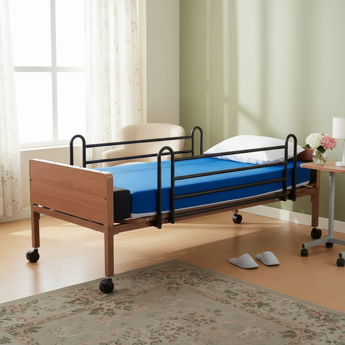 A hospital bed with black metal side rails and a Protekt Mattress Extender installed at the foot end, lengthening the blue-covered mattress surface. The bed is in a room with a window and a rug.