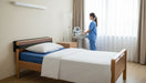 A hospital bed set up with a Protekt Wall Guard attached to the wooden headboard, protecting the wall behind it. A nurse in blue scrubs is visible in the background near a window.