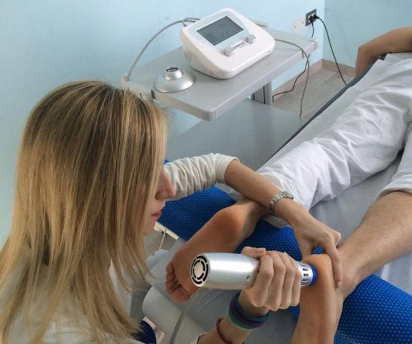 A clinician applying shockwave therapy to the patient's foot/heel area using the Oceanus PhysioPRO shockwave device handle, with the control unit visible in the background.