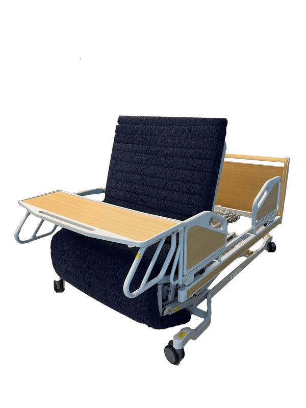 The ReNuCare Standing Bed is a 4-position adjustable hospital bed with metal rails, wood headboard and footboard, and tray table. Set upright on a wood-patterned floor, it offers enhanced comfort and support.