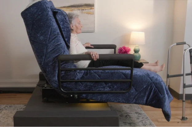 An elderly woman reclines comfortably on a UPbed Independence Adjustable Bed for Seniors by Journey Health, featuring blue bedding and side rails. Nearby are a walker, lamp, and flowers, highlighting safety and a supportive living environment.
